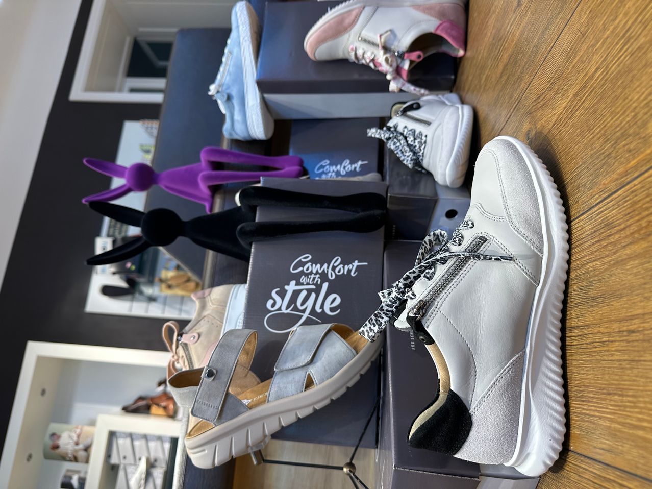 A wooden table displays various shoes, including sneakers, sandals, and loafers, with the brand name 'Comfort with style' on the boxes.