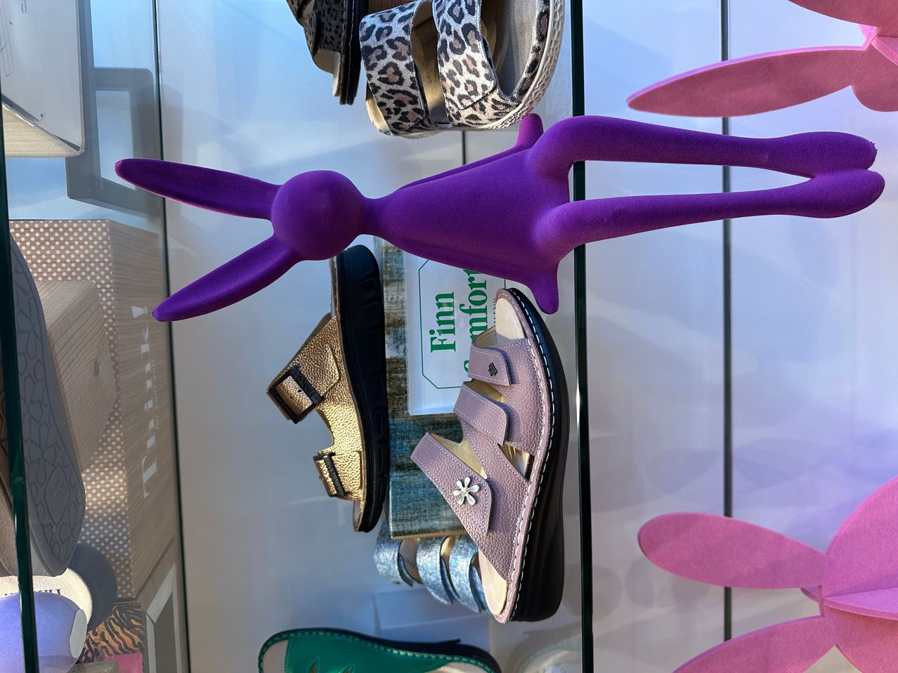 A purple toy rabbit stands above a display of sandals in a shop window. The sandals are in various colors and styles, with a sign that reads 'Finn comfort.'