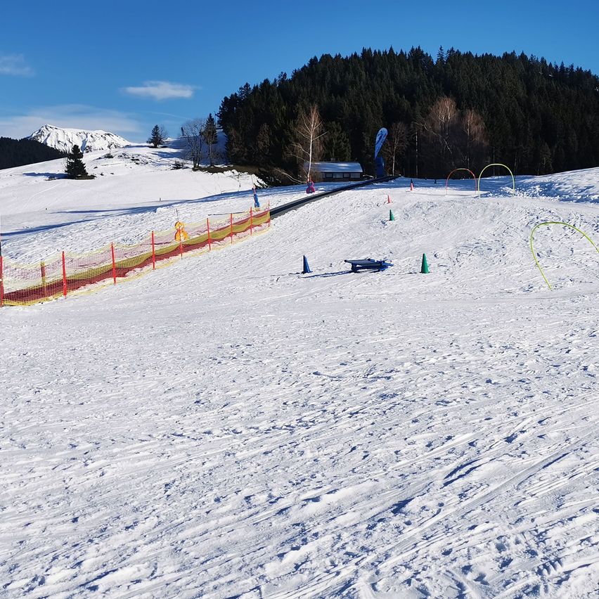 Eine verschneite Landschaft mit klarem blauem Himmel, Bäumen und Bergen im Hintergrund. Im Vordergrund befindet sich ein schneebedeckter Hang mit einer Umzäunung, mehreren Hindernissen und einer blauen Bank.