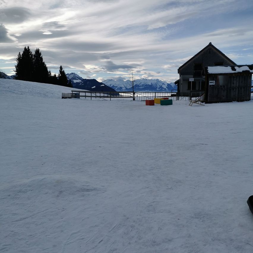 Eine verschneite Landschaft mit einem kleinen Holzgebäude, möglicherweise einer Skihütte, umgeben von Bäumen und Bergen in der Ferne. Der Himmel ist bewölkt.