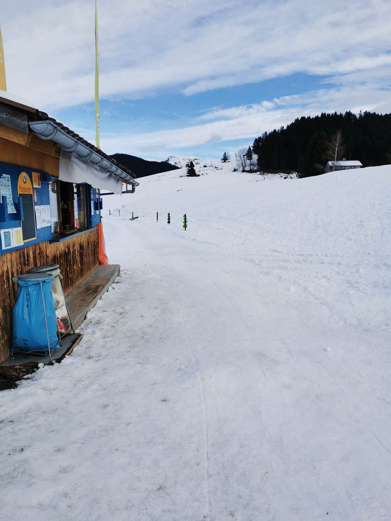 Eine Skihütte auf einem verschneiten Berghang. Die Hütte hat ein hölzerne Außenseite und blaue Schilder. Der Schnee ist dick und weiß, mit Reifenspuren und Kiefern im Hintergrund.