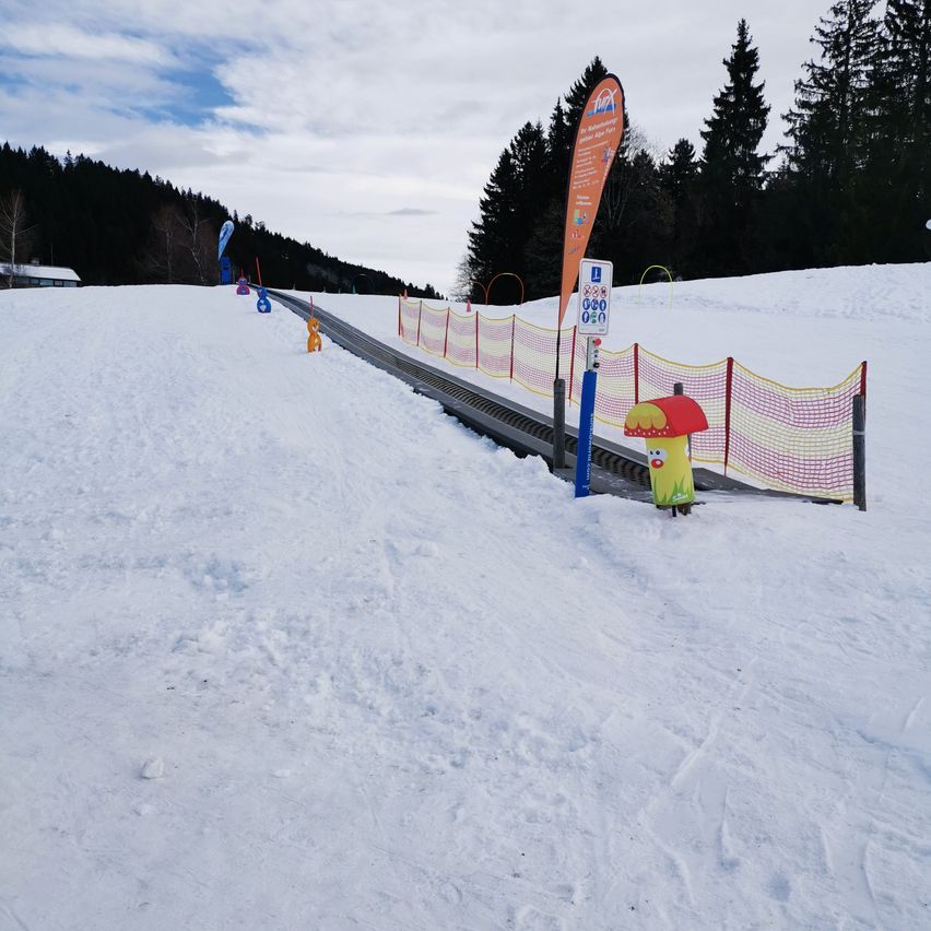 Eine verschneite Landschaft mit einem gewundenen Pfad, einem bunten Pilz und einem Zaun. In der Ferne befinden sich Bäume und ein Gebäude.