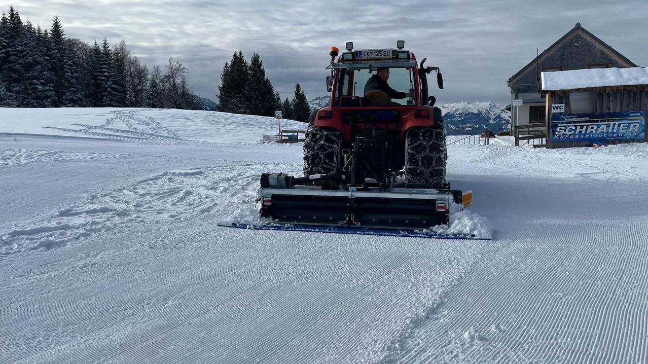 Ein roter Traktor mit einem Schneepflug fährt durch ein verschneites Feld. Ein Mann bedient den Traktor, dessen Kennzeichen FK 125 EU lautet. Die Szene ist vor dem Hintergrund von Bergen und Bäumen angesiedelt.