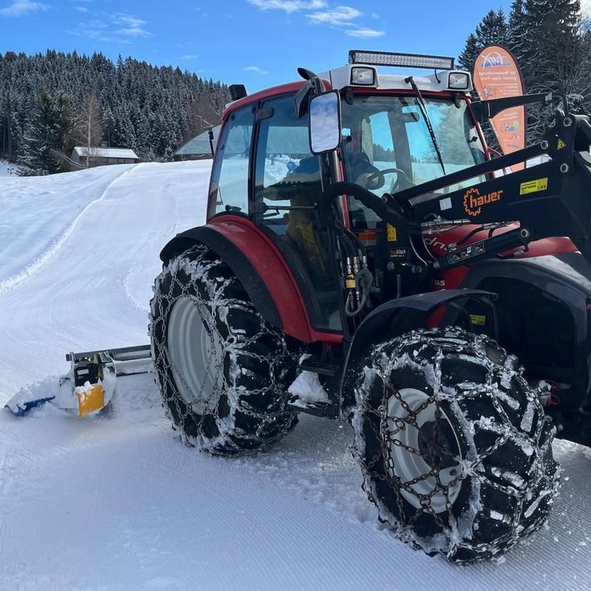 Ein roter Traktor mit Ketten an den Reifen pflugt eine schneebedeckte Straße.
