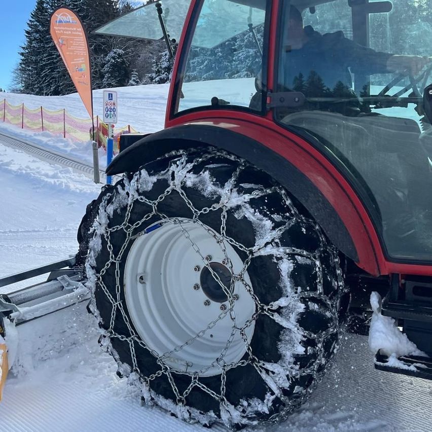 Ein roter Traktor mit Ketten an den Reifen fährt auf einem verschneiten Pfad. Dahinter befinden sich eine Fahne und ein Schild. Der Traktorfahrer trägt eine Jacke und Handschuhe.