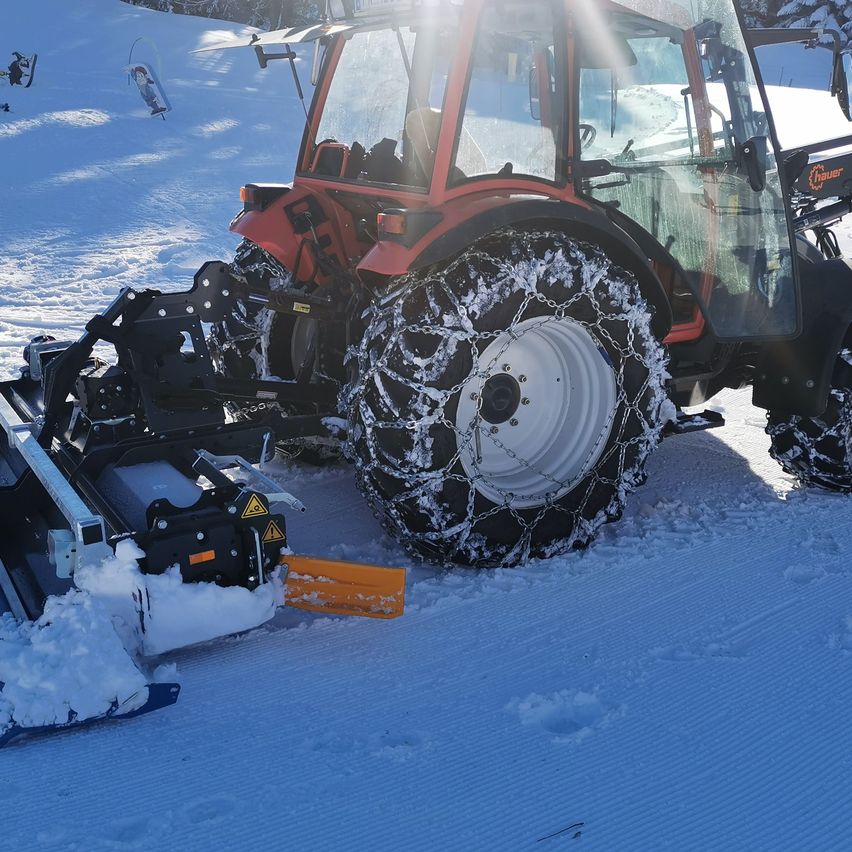 Ein roter Traktor mit Schneeketten pflügt einen verschneiten Pfad, mit Bäumen und Menschen im Hintergrund.
