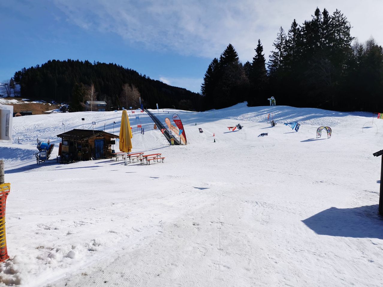 A ski resort with snow-covered slopes, a small building, and several picnic tables. Trees surround the area, and a skier is visible in the distance.