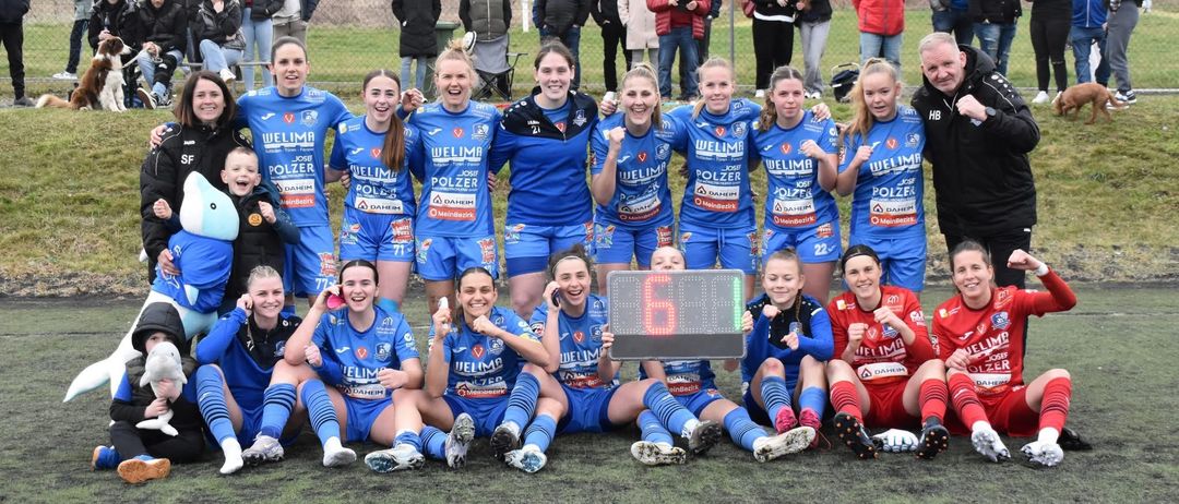 Frauen-Fußballteam in blauen Trikots posiert für ein Foto auf einem Feld mit einem digitalen Punktestand von 60, mit Zuschauern im Hintergrund.
