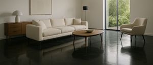 Ein minimalistisches Wohnzimmer mit einer weißen Couch, einem runden Holztisch und großen Glastüren, die in einen Garten führen. Ein gerahmtes Bild und eine schwarze Lampe sind ebenfalls vorhanden.