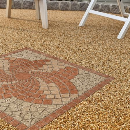 Ein dekoratives quadratisches Steinmosaik mit Spiralmuster auf einer Kiesoberfläche, umgeben von zwei weißen Stühlen.