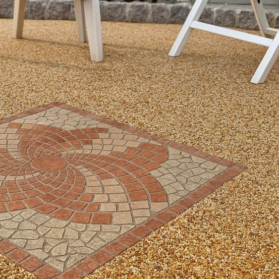 Ein dekoratives quadratisches Steinmosaik mit Spiralmuster auf einer Kiesoberfläche, umgeben von zwei weißen Stühlen.