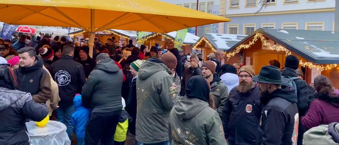 Eine große Menschenmenge versammelt sich auf einem Weihnachtsmarkt unter einem gelben Schirm, mit festlich geschmückten Ständen und einem großen Gebäude im Hintergrund.