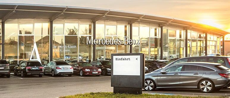 Mercedes-Benz Autohaus mit mehreren geparkten Autos davor, darunter ein graues Auto vorne. Die Sonne geht unter.