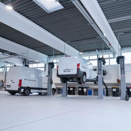 Ein weißer Van wird in einer Garage von einem Hebebühne angehoben. Ein weiterer Van steht dahinter. Zwei Autolifte sind mit 'MA STAAR' beschriftet.
