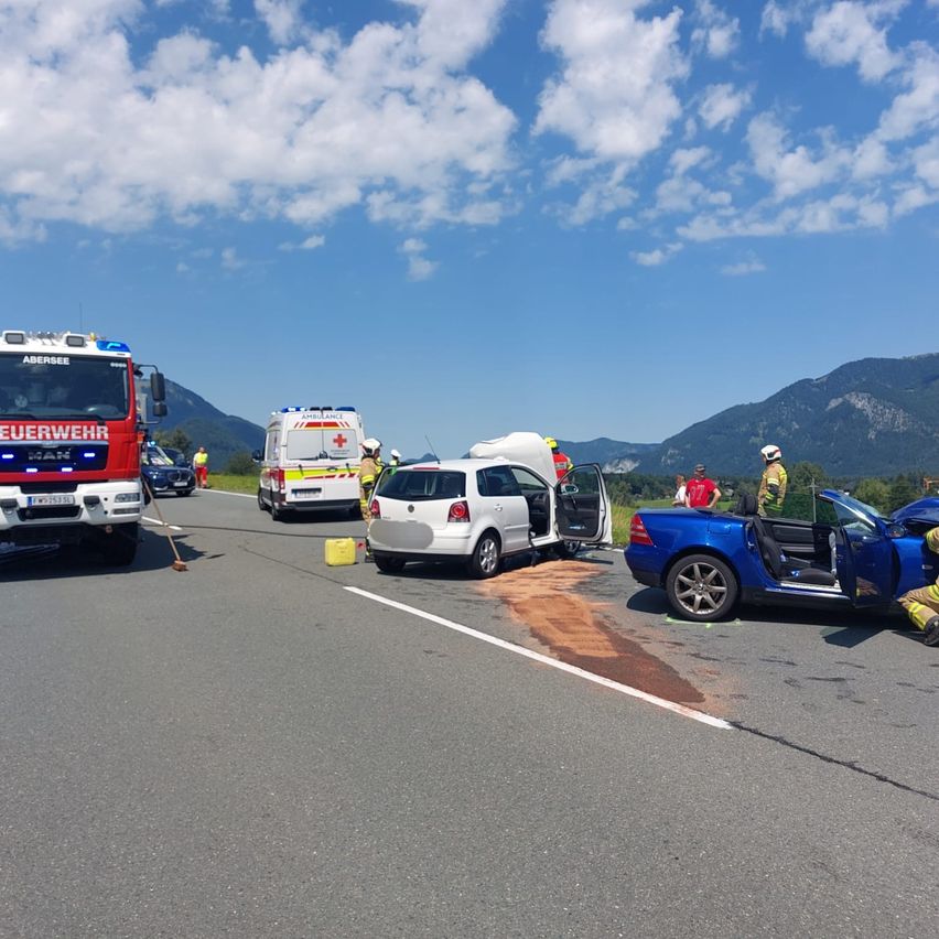 Rettungsfahrzeuge und Personal an einem Unfall auf der Straße. Ein roter Feuerwehrwagen, ein Krankenwagen und ein blaues Auto sind beteiligt. Menschen stehen herum, und die Szene befindet sich auf einer Straße mit Bergen im Hintergrund.
