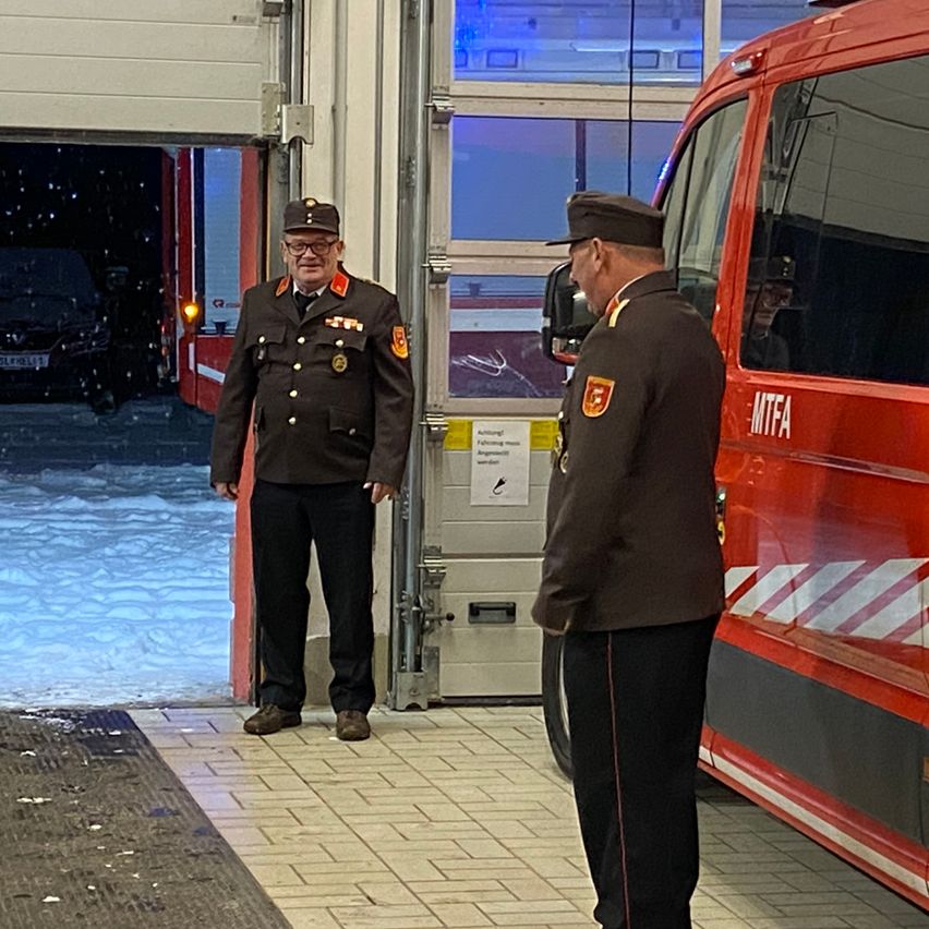Zwei Männer in Uniform stehen vor einem roten Feuerwehrauto. Sie sind drinnen, wahrscheinlich in einer Garage. Schnee ist auf dem Boden draußen sichtbar.