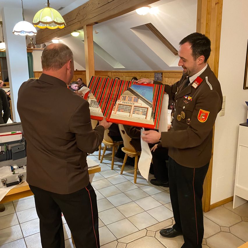 Zwei Männer in Militäruniformen halten eine verpackte Box in einem Raum mit Holzbalken, während andere auf Stühlen sitzen. Ein Schrank, eine Lampe und ein gefliester Boden sind sichtbar.