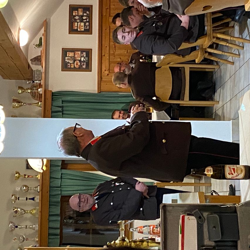 Mehrere Personen in Uniform sitzen in Stühlen in einem Raum mit Trophäen an der Wand. Ein Mann steht vor ihnen und blickt nach unten. Eine Trophäe steht auf dem Tisch.