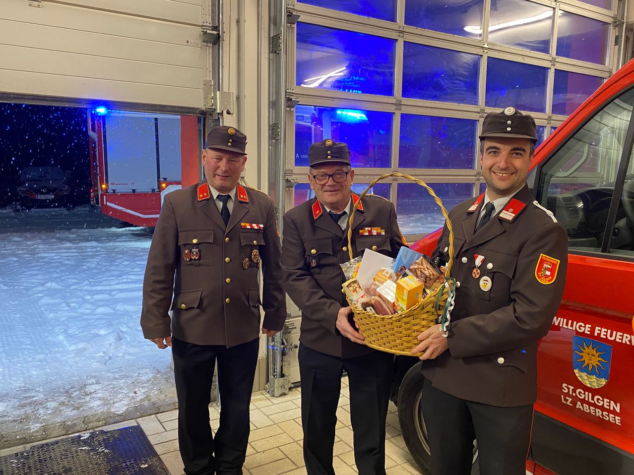 Drei Feuerwehrleute in Uniform stehen in einer Garage. Ein Mann hält einen Korb mit Lebensmitteln.