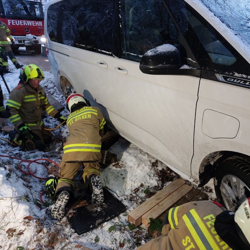 Firefighters in protective gear are rescuing a van stuck in snow. One person kneels beneath the vehicle while another stands nearby. A fire truck is parked behind them.