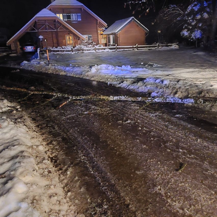 A night scene shows a snowy driveway leading to a log cabin with a car parked under a garage. Snow covers the ground and a fence borders the property.