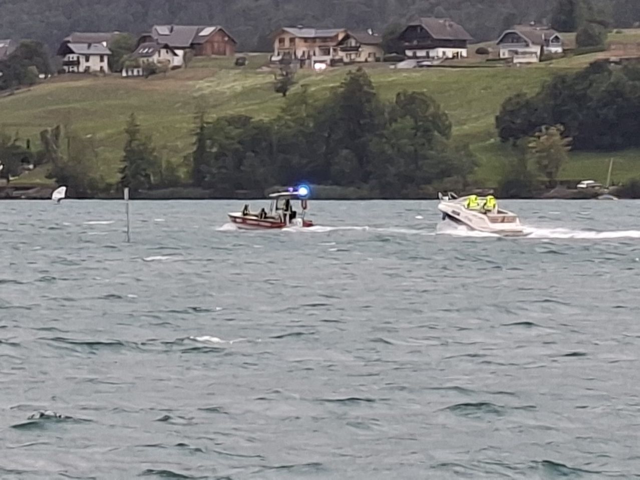 Zwei Boote sind auf einem See mit blinkenden Lichtern, eines vor dem anderen, in der Nähe eines Hügels mit Häusern.