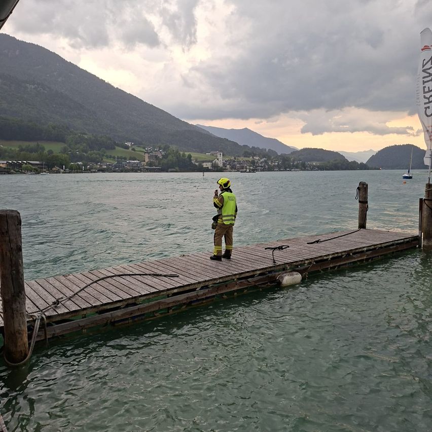 Eine Person in einer gelben Sicherheitsweste steht auf einem Holzsteg am Wasser, mit Bergen und einem bewölkten Himmel im Hintergrund.
