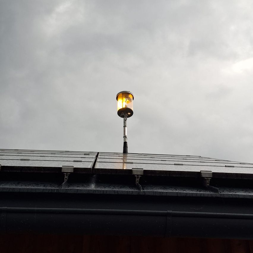 Eine solarbetriebene Laterne ist auf einem Dach unter einem bewölkten Himmel montiert. Die Laterne ist beleuchtet und strahlt ein warmes Licht aus.