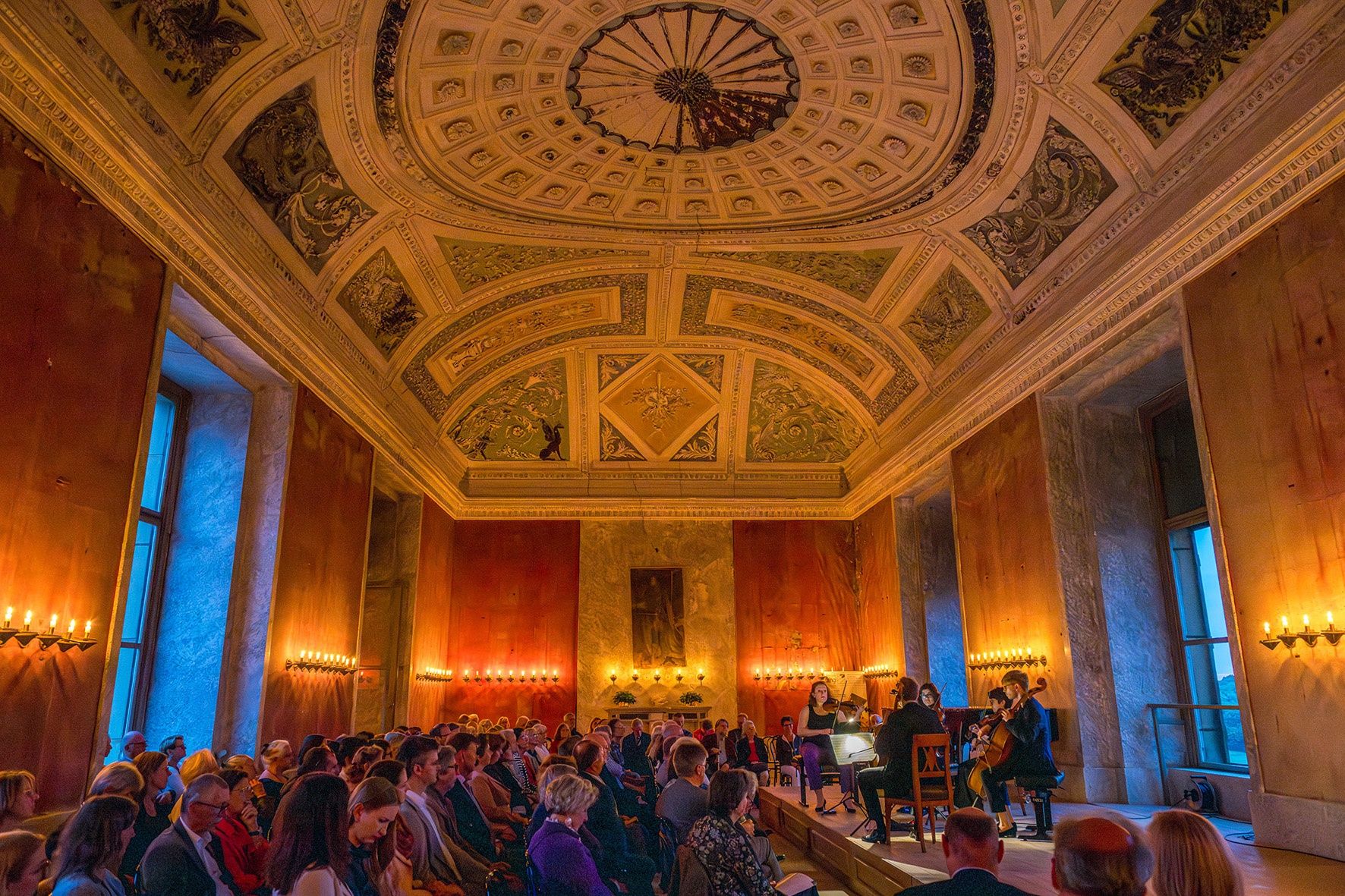 Ein elegantes Kammermusikensemble spielt auf der Bühne mit Streichinstrumenten in einem historischen Saal mit verzierten Wänden und einer hohen, dekorativen Decke. Das Publikum sitzt aufmerksam in Reihen, und der Raum wird sanft von Kronleuchtern beleuchtet.