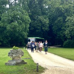 Eine Gruppe von Menschen geht einen Kiesweg entlang, mit einem Bus im Hintergrund, umgeben von Bäumen und Gras.