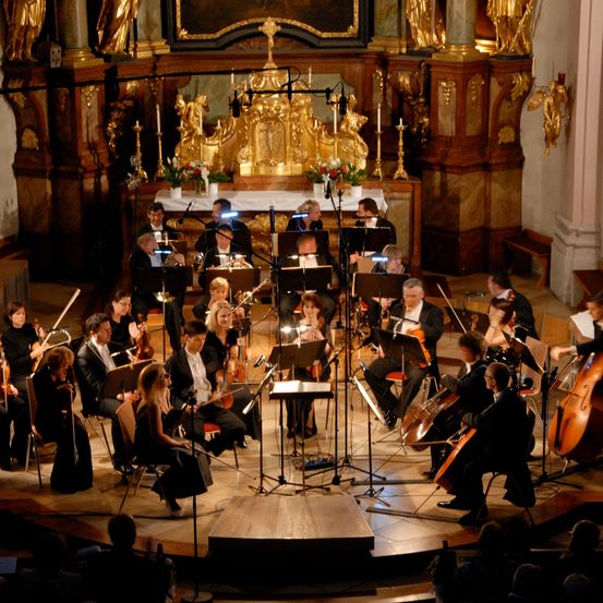 Ein Orchester spielt auf einer Bühne in einer Kirche mit einem Altar und goldenen Dekorationen im Hintergrund. Die Musiker spielen Violinen, Celli und andere Instrumente.