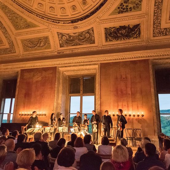 Ein klassisches Musikensemble spielt auf der Bühne mit Violinen und Celli, während ein Publikum von unten zuschaut. Der Raum hat große Fenster und dekorative Deckendesigns.