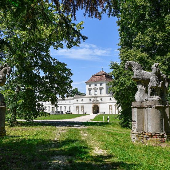 Ein großes weißes Schloss mit einem braunen Dach und großen Fenstern steht in einem Garten. Zwei Steinpferdestatuen befinden sich auf beiden Seiten. Der Garten ist üppig mit grünem Gras.