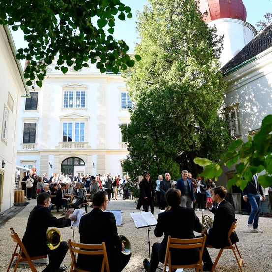 Eine Gruppe Musiker spielt auf dem Außenhof eines historischen Gebäudes, das von vielen Menschen beobachtet wird. Das Gebäude hat eine rote Kuppel und große Fenster. Bäume und Pflanzen umgeben das Gelände.