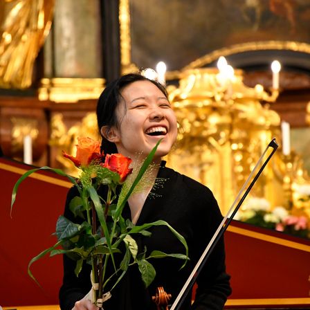 Eine Frau hält einen Strauß Rosen, lächelt breit, steht in einer Kirche mit goldenem Hintergrund und hält eine Geige.