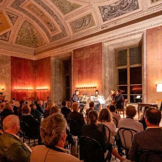 Ein klassisches Musikkonzert findet in einem wunderschön dekorierten Raum statt. Die Musiker spielen Violinen, und das Publikum verfolgt aufmerksam die Aufführung. Der Raum hat kunstvoll verzierte Decken und Wände mit Kerzenständern.