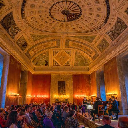 Eine Gruppe Musiker tritt auf der Bühne in einem großen Saal mit kunstvollen Verzierungen und Kerzenlicht auf, während ein Publikum aufmerksam zuschaut.