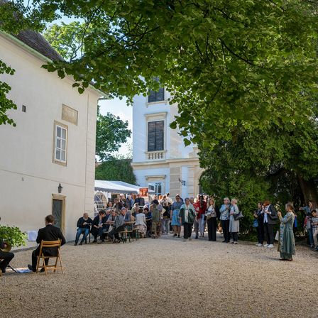 Eine Gruppe von Menschen versammelt sich vor einem Gebäude, einige sitzend und andere stehend. Ein Mann spielt ein Musikinstrument auf einem Stuhl. Bäume und Pflanzen sind vorhanden.