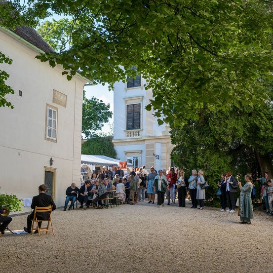 Eine Gruppe von Menschen versammelt sich vor einem Gebäude, einige sitzend und andere stehend. Ein Mann spielt ein Musikinstrument auf einem Stuhl. Bäume und Pflanzen sind vorhanden.
