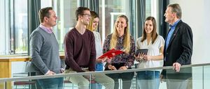 Fünf Personen unterhalten sich in einem modernen Büro. Ein Mann in einem weinroten Hemd sitzt, während eine Frau ein Tablet hält. Zwei Frauen stehen, eine mit einer roten Mappe.