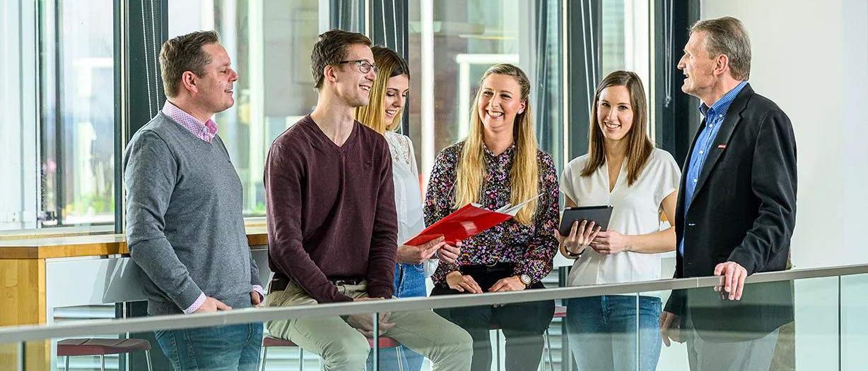 Fünf Personen unterhalten sich in einem modernen Büro. Ein Mann in einem weinroten Hemd sitzt, während eine Frau ein Tablet hält. Zwei Frauen stehen, eine mit einer roten Mappe.