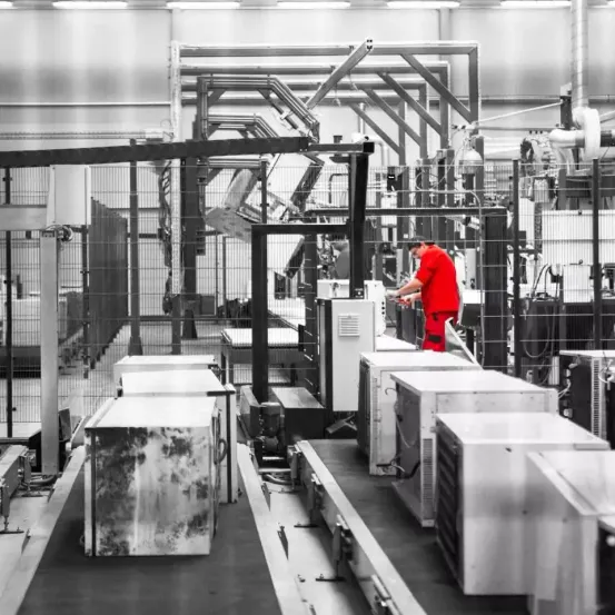 Ein Schwarz-Weiß-Bild einer Fabrik mit einem Arbeiter in einem roten Shirt, der Maschinen inspiziert. Industriegeräte und Förderbänder sind sichtbar.