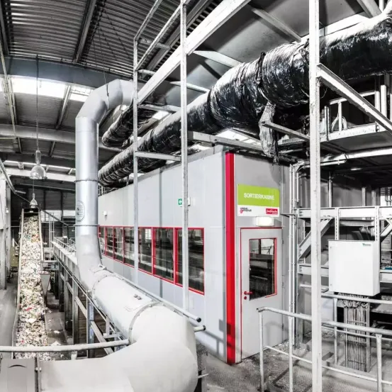 Ein Fabrikinnenraum mit großen Rohren, Metallstrukturen und Maschinen. Ein Förderband auf der linken Seite transportiert Abfallmaterialien. Ein Gebäude mit roter Tür und grünem Schild befindet sich auf der rechten Seite.