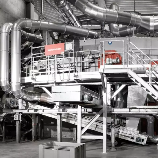 Ein Schwarz-Weiß-Bild einer Industrieanlage mit Metallrohren und -strukturen, einem roten Schild mit dem Wort 'Windsichter' und einem großen Metallcontainer unten.