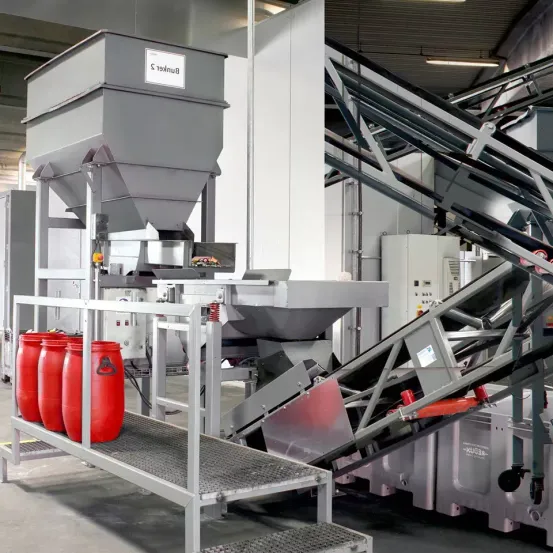 Eine Fabrik mit einer großen Industriemaschine. Eine Metallplattform hält vier rote Fässer, wobei ein Förderband zu einer größeren Maschine führt.