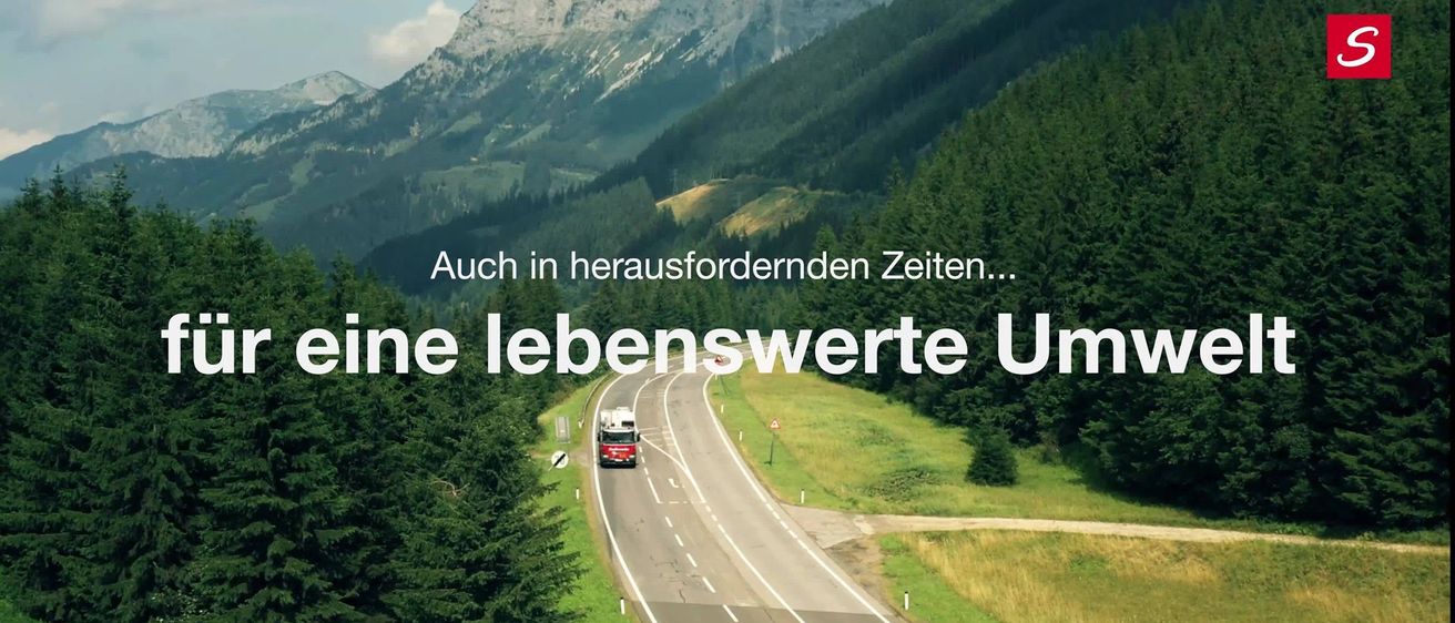 Eine Autobahn mit einem fahrenden LKW, eingerahmt von Bergen und Bäumen. Der Text überlagert lautet 'Auch in herausfordernden Zeiten...' mit einem motivierenden Satz darunter.