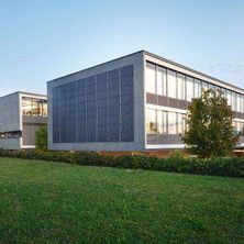 Ein modernes Bürogebäude mit Solarpaneelen an der Fassade, eingebettet in eine grüne Landschaft mit Bäumen und Gras.