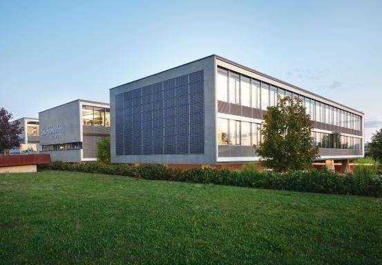 Ein modernes Bürogebäude mit Solarpaneelen an der Fassade, eingebettet in eine grüne Landschaft mit Bäumen und Gras.