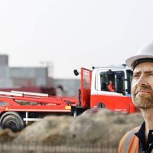 Ein Bauarbeiter steht vor einem roten Maschinenfahrzeug, trägt einen weißen Helm und eine orange Weste. Das Maschinenfahrzeug ist hinter ihm geparkt, und eine Person sitzt drinnen.
