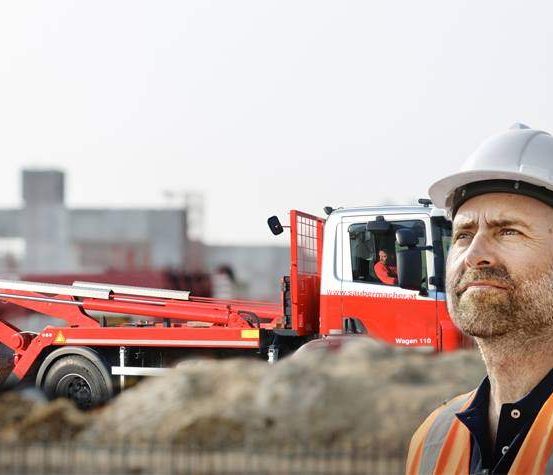Ein Bauarbeiter steht vor einem roten Maschinenfahrzeug, trägt einen weißen Helm und eine orange Weste. Das Maschinenfahrzeug ist hinter ihm geparkt, und eine Person sitzt drinnen.